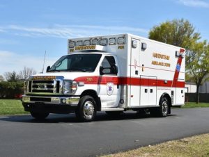 A white ambulance with red stripes and the text "HAVERSTRAW AMBULANCE CORPS" on the side and front, parked on a paved road near grassy areas and trees under a clear sky.