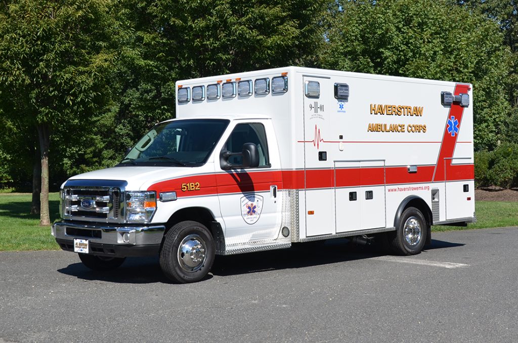 A white and red Haverstraw Ambulance Corps ambulance is parked on a paved area surrounded by trees and greenery. The vehicle has a blue star of life symbol and various medical and emergency markings on its sides.