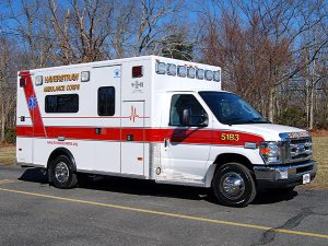 A white and red ambulance with "Haverstraw Ambulance Corps" written on the side is parked on a paved road against a backdrop of leafless trees. The vehicle has emergency medical symbols and numbers displayed, and a website URL is visible on the lower side.