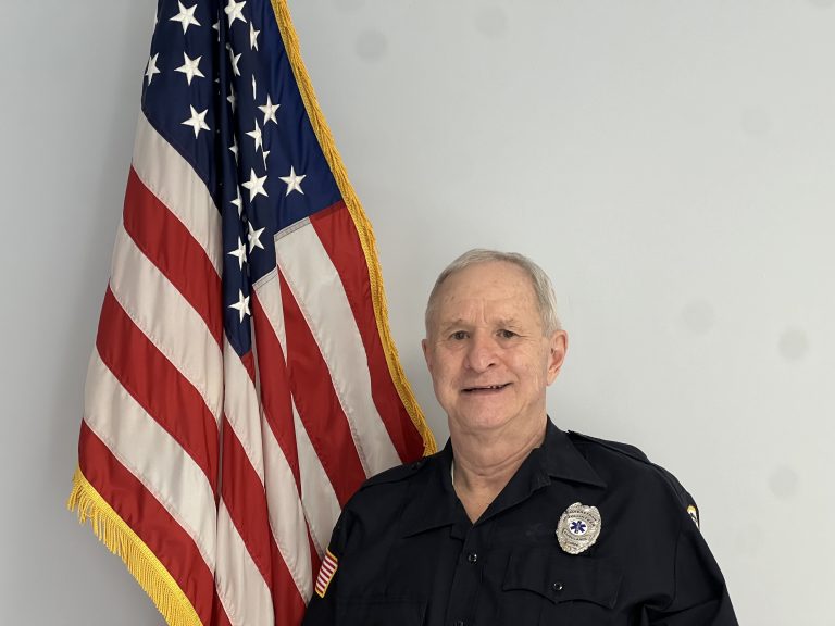 An older man in a dark uniform with a badge stands smiling in front of a U.S. flag.