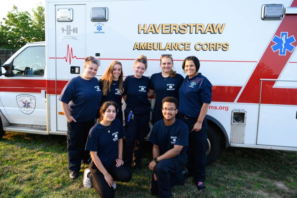 A group of seven people in dark blue uniforms stand in front of a Haverstraw Ambulance Corps vehicle. They are smiling and posing on a grassy area, with trees visible in the background.