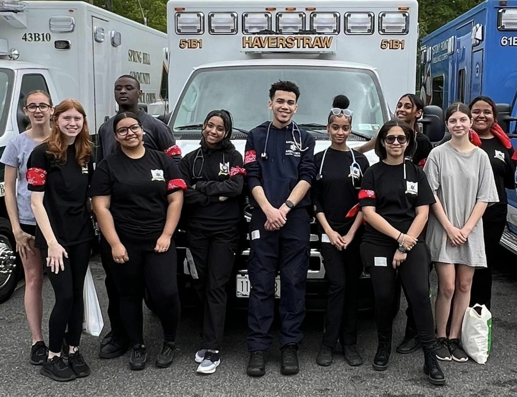 A group of people in black uniforms stand together in front of an ambulance that reads "Haverstraw." Some are wearing stethoscopes, and they all smile at the camera. Two other emergency vehicles are parked nearby.