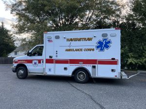 A white and red ambulance with "Haverstraw Ambulance Corps" written on the side is parked on a road. An American flag and medical star emblem are also visible. Trees and a wooden fence are in the background.