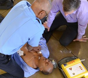 Two men assist another on the floor with an AED. One man performs CPR chest compressions, while the other holds the patient's head. An AED device is visible on the ground nearby.